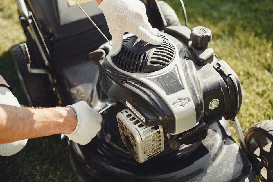 Close-up view of a person's gloved hands operating a gas-powered lawn mower on a well-maintained garden lawn. The mower features a black and silver engine with visible air vents and control components