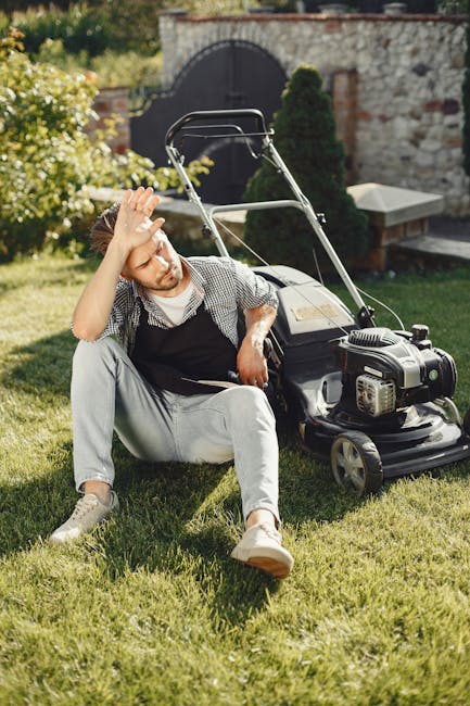 Close-up view of a person's gloved hands operating a gas-powered lawn mower on a well-maintained garden lawn. The mower features a black and silver engine with visible air vents and control components