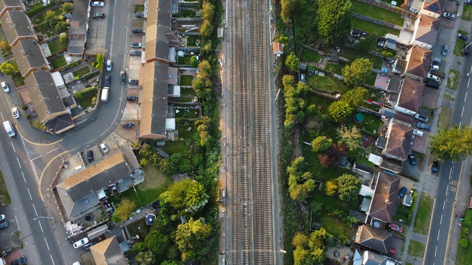 An aerial view showing a narrow strip of land with railway tracks running through the center, bordered by residential gardens and houses on both sides. To the left, there is a curved road with parked 